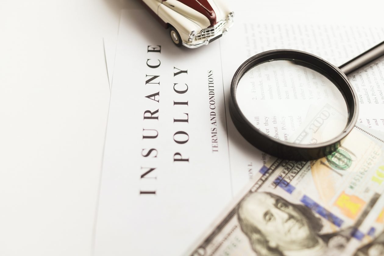 Image of document titled "Insurance Policy" with money, car, and magnifying glass sitting on top of the document.